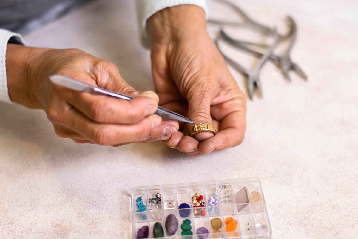 Artisan bijoutier travaillant sur une bague en or avec un outil de précision, entouré de pierres précieuses et de matériel de joaillerie.